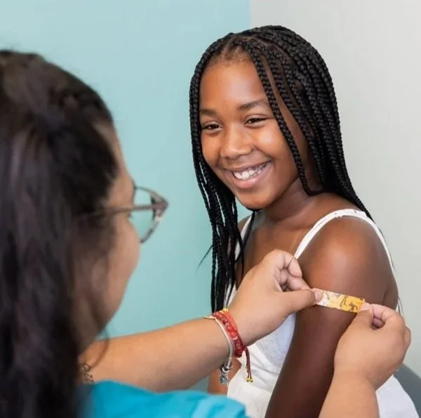 A young girl with braided hair smiling as a healthcare worker administers a vaccine to her upper arm.