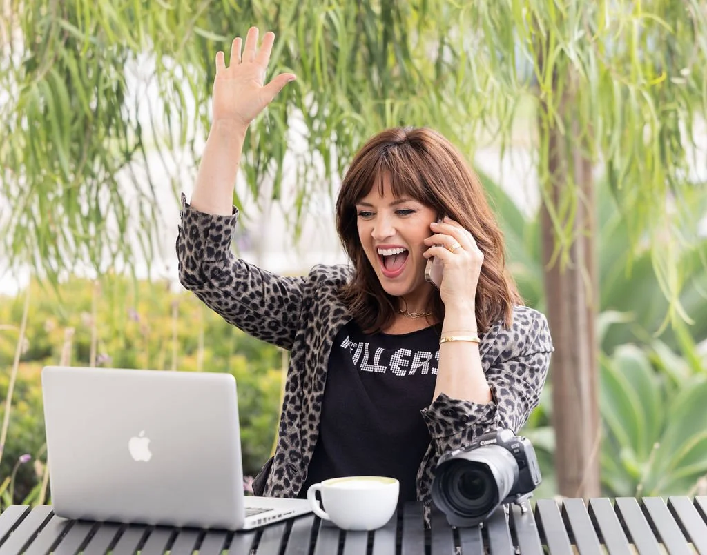 A woman with brown hair and a leopard print jacket, sitting at an outdoor table, smiling and talking on her phone while waving. On the table are a silver laptop, a camera, and a white coffee cup. The background features green plants and trees.