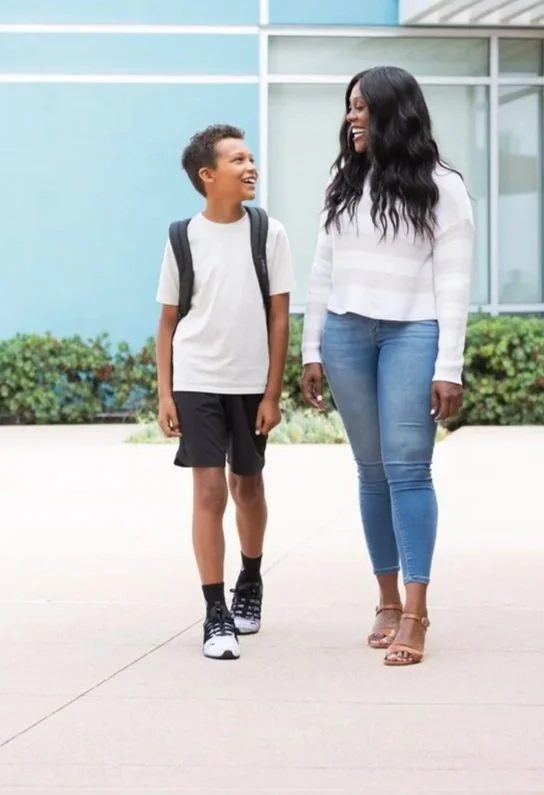 A woman and a boy walking outdoors and smiling at each other, with a building and some bushes in the background.