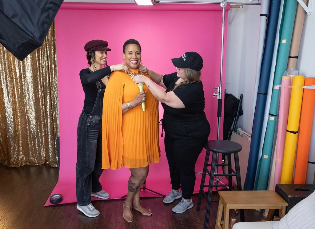 Three women in a photography studio, with one woman wearing an orange dress and holding a gold microphone. The other two women are adjusting jewelry and accessories on her, smiling. The studio has a pink backdrop, gold curtain, and colorful rolls of backdrop paper.