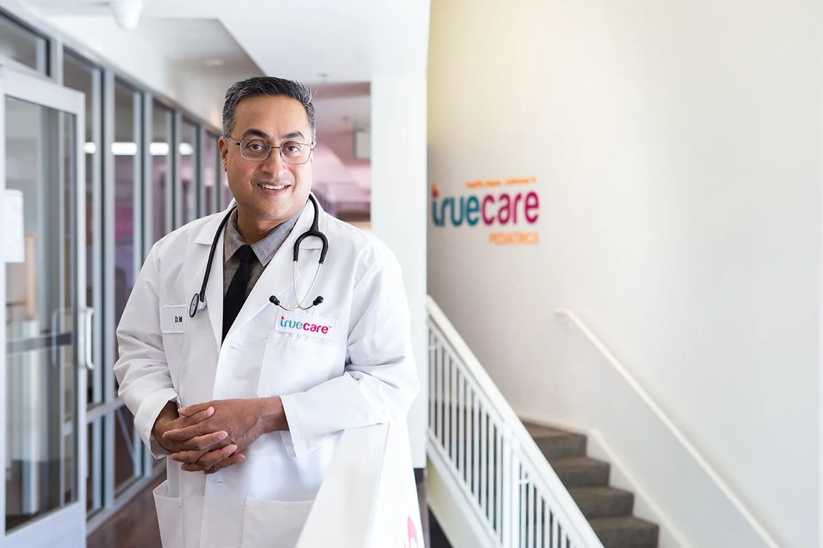 A male doctor standing in a healthcare facility, smiling and wearing a white coat and stethoscope, in front of a sign that says 'true care'.