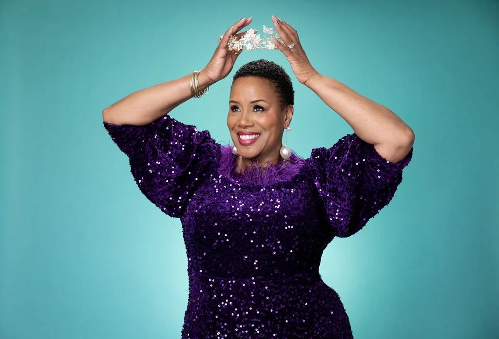 A woman wearing a purple sequined dress, holding a tiara above her head, smiling, set against a teal background.