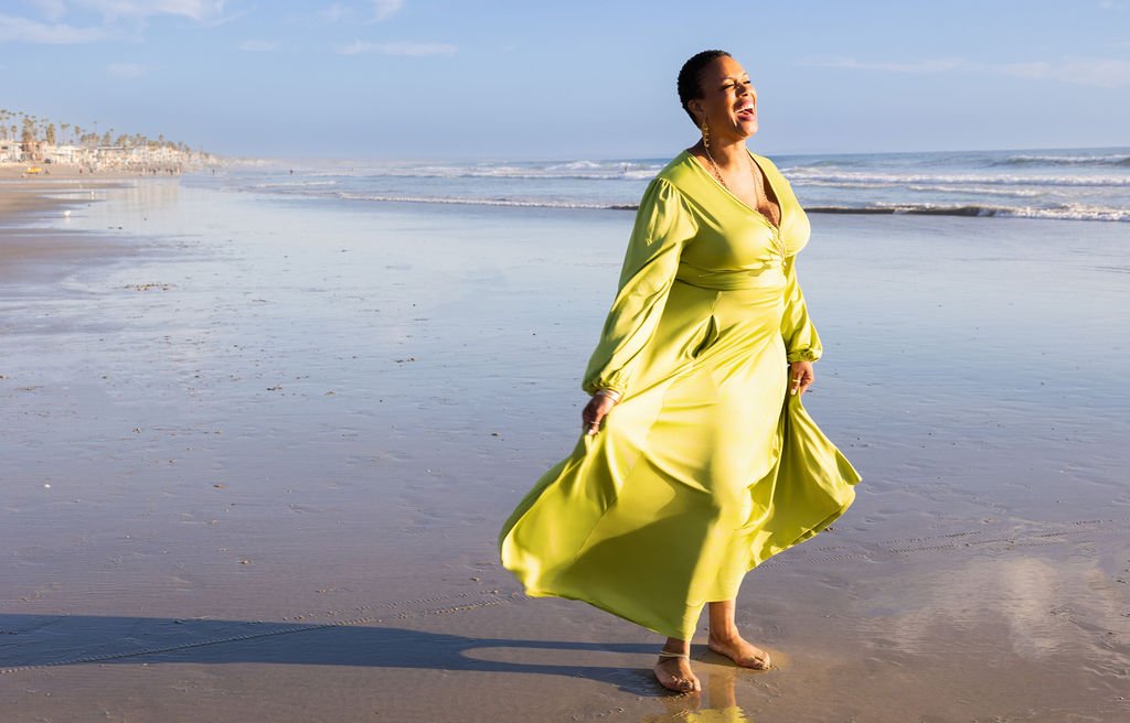 A woman in a bright yellow dress standing barefoot on the beach near the water with ocean waves in the background, smiling.
