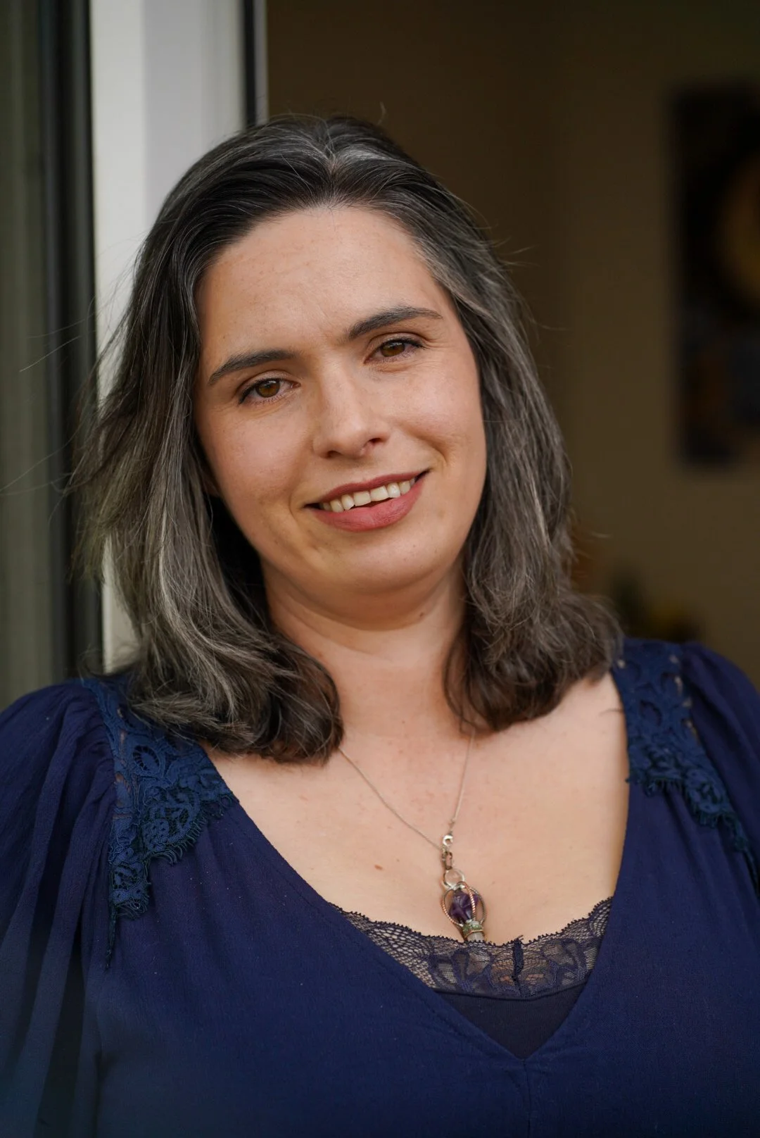 A woman with shoulder-length gray and brown hair, smiling, wearing a navy blue top with lace details and a necklace with a purple gemstone pendant, standing near a door.
