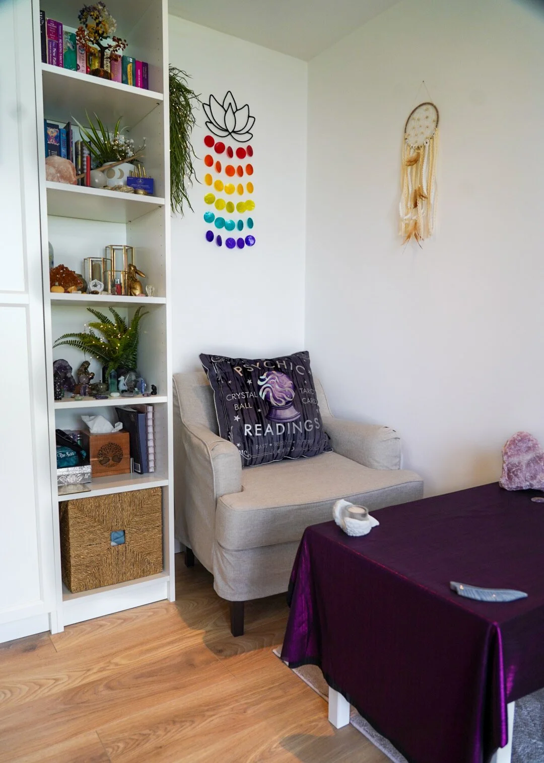 Interior of a cozy room with a white bookshelf, a beige armchair with a psychic readings pillow, a purple cloth-covered table, and wall decorations including a rainbow-colored chakra wall hanging and a dreamcatcher.