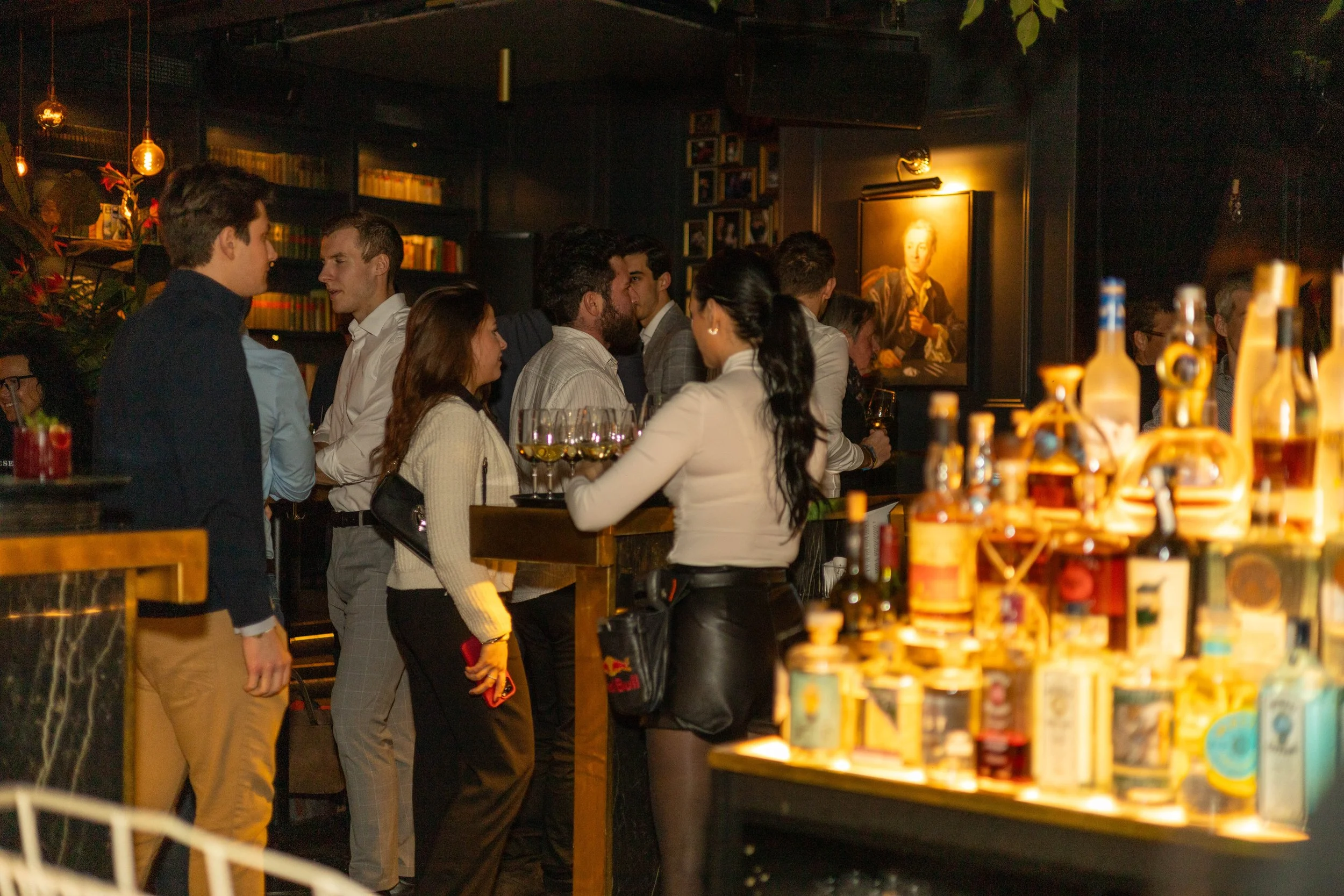 Menschen stehen in einer Bar oder einem Restaurant bei Nacht. Sie unterhalten sich, einige halten Weingläser. Im Hintergrund hängt ein Porträt an der Wand, und im Vordergrund sind Spirituosenflaschen auf einem Regal sichtbar.
