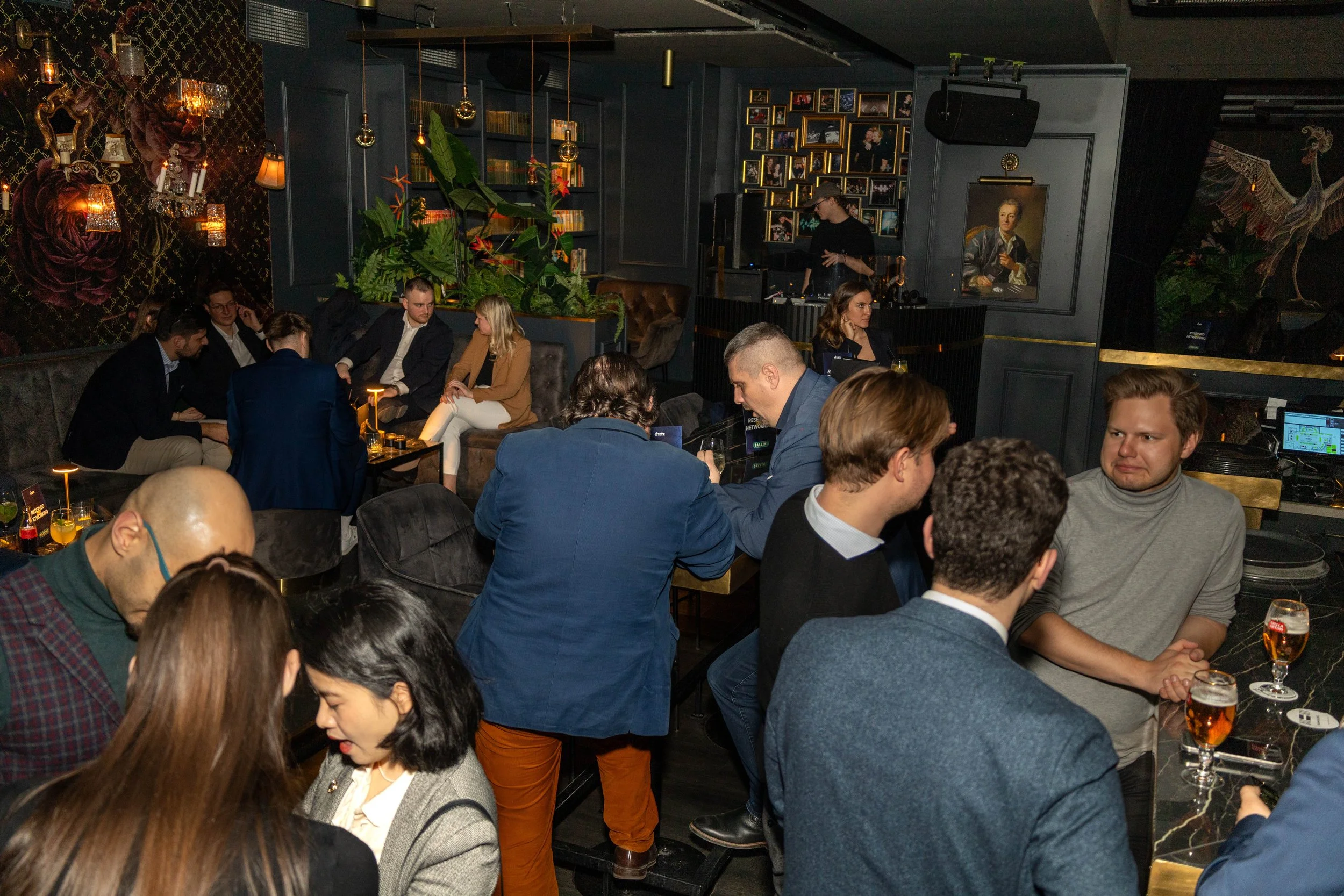 Menschen sitzen und stehen in einer dunklen, eleganten Bar, während ein DJ im Hintergrund auflegt. Der Raum ist mit Pflanzen, Fotos und dekorativen Elementen geschmückt, die eine stilvolle Atmosphäre schaffen.