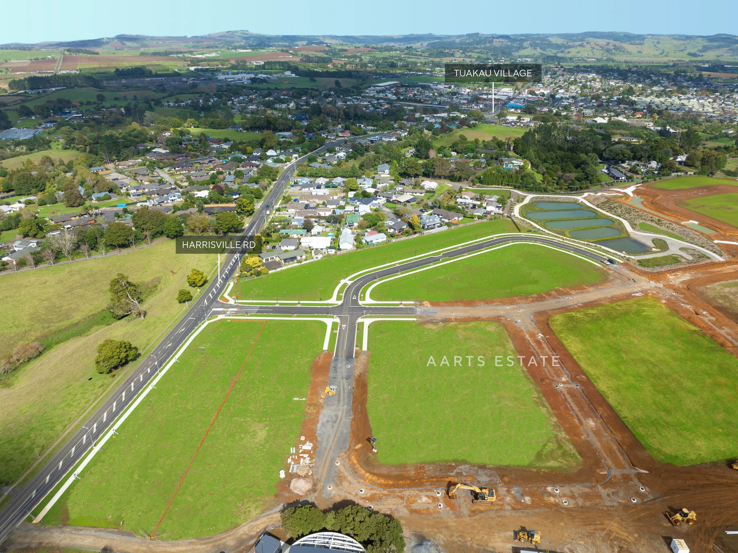 Aerial view of a suburban neighborhood under construction, with roads and houses, and a section of land being developed with construction equipment and pathways, near Tuakau Village.