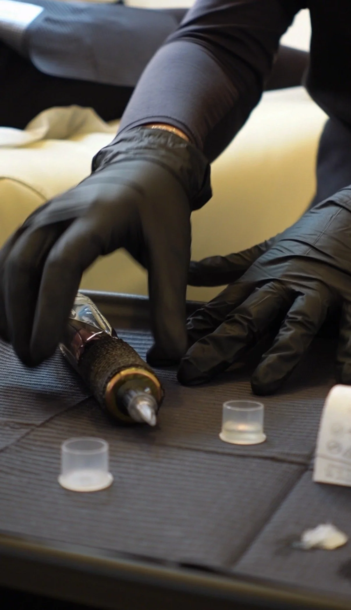 Person wearing black gloves working with a syringe and small vials on a textured surface.