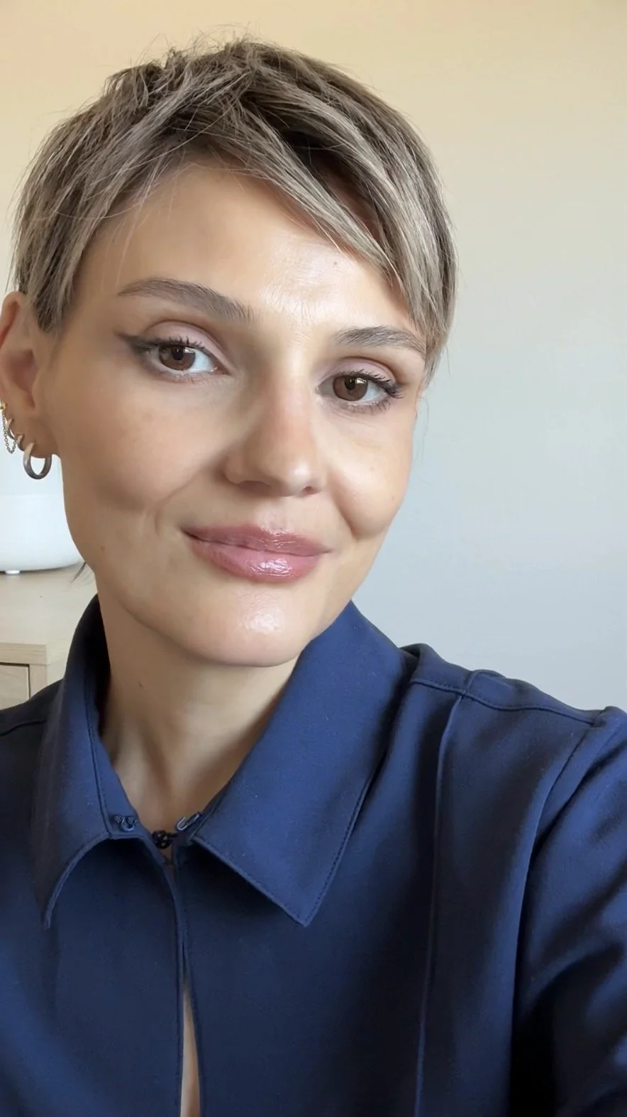 A woman with short, light brown hair styled in a pixie cut, wearing a dark blue collared shirt, smiling softly, with earrings in her left ear, in an indoor setting with a white wall and a wooden side table visible in the background.