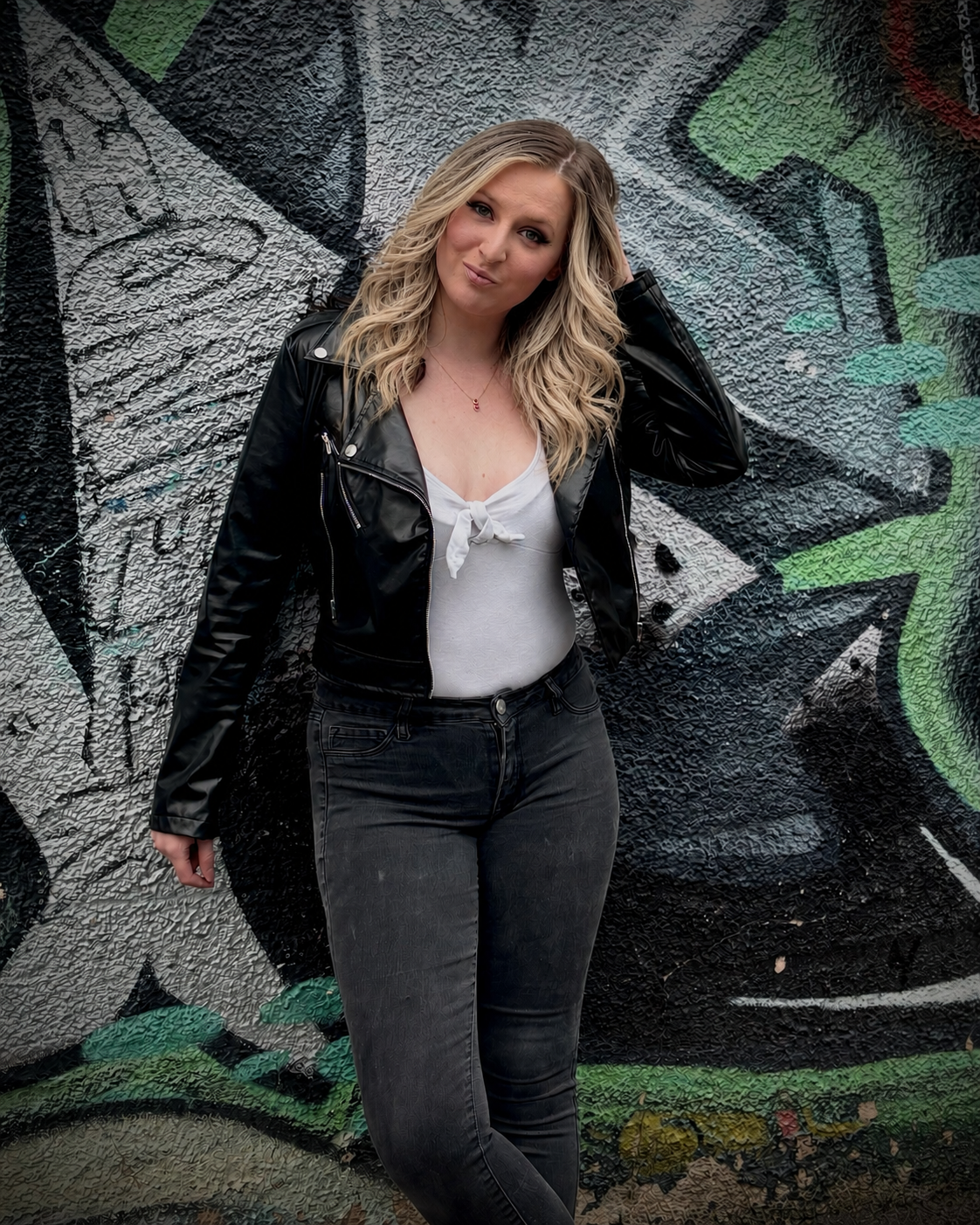 Young woman with wavy blonde hair wearing a black leather jacket, white top, and black jeans, standing in front of a graffiti-covered wall.