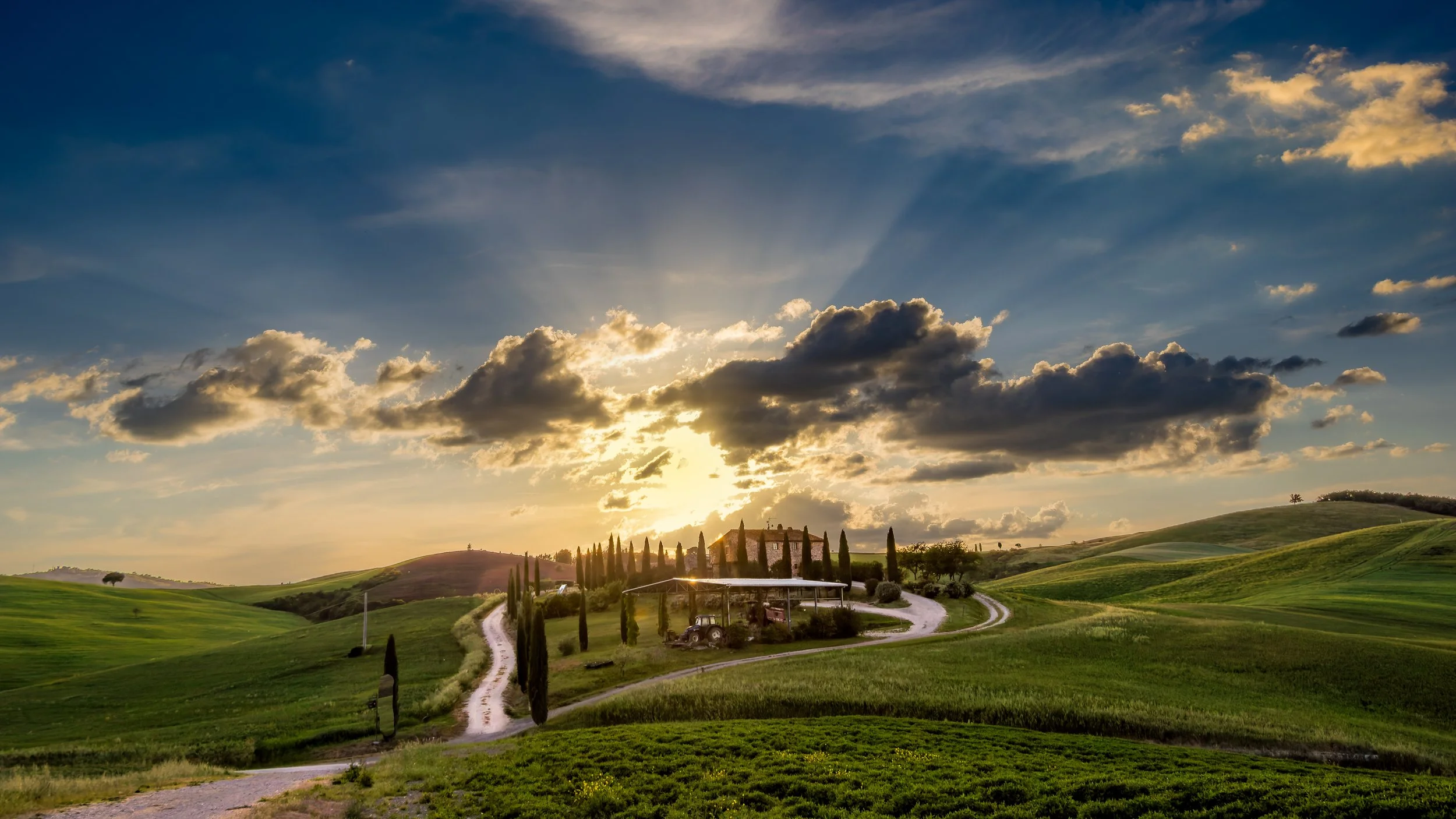 Sunset over rolling green hills with cypress trees, a winding dirt road, and a farmhouse in the distance under partly cloudy sky.