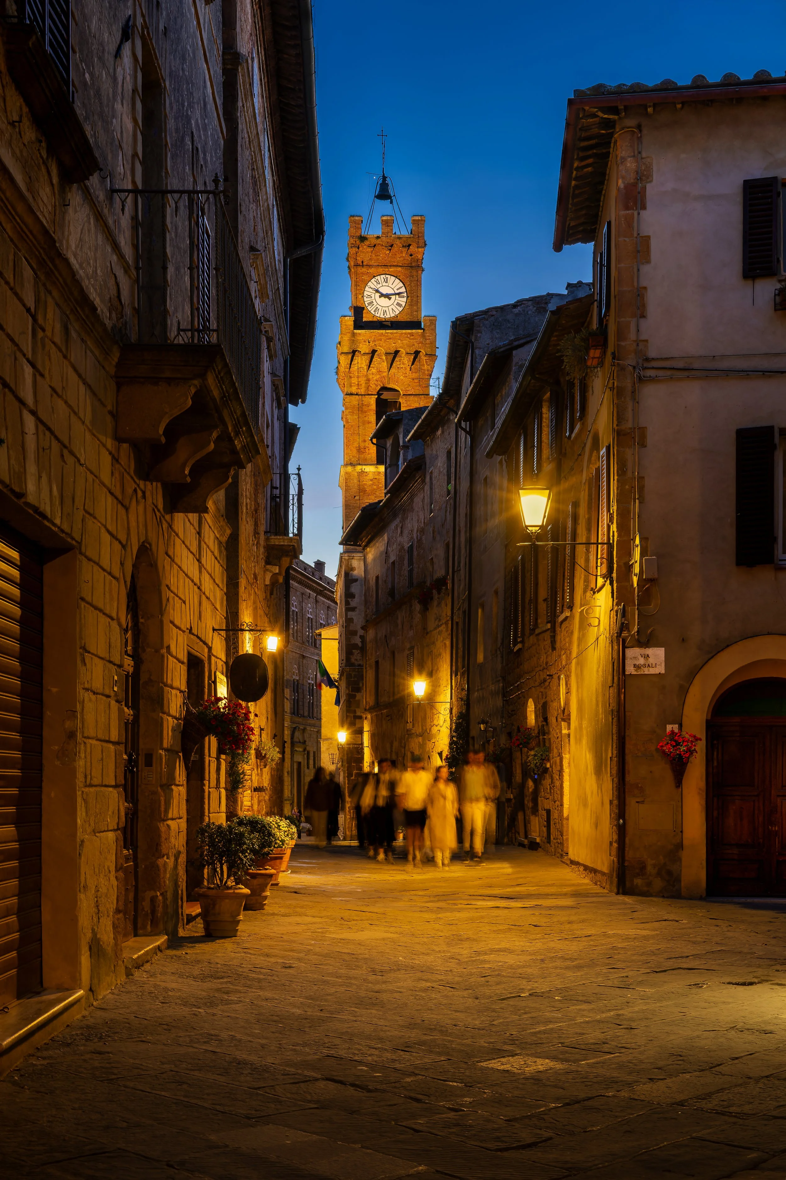 A narrow cobblestone street in an Italian town, Pienza, during dusk, illuminated by street lamps. There are a few people walking, and an old clock tower is visible in the background against a deep blue sky.