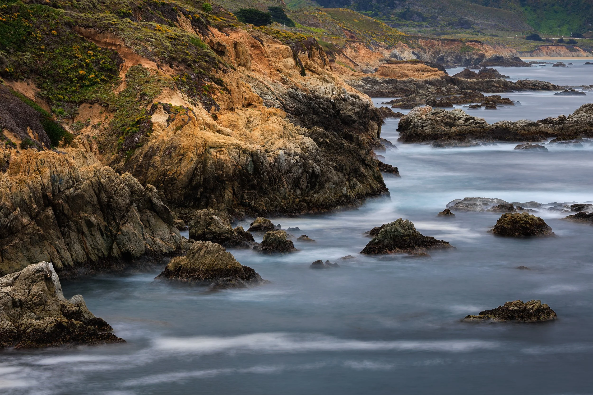 A rugged coastline with large rocks and cliffs covered in green bushes, ocean waves crashing against the rocks, and distant hills in the background under a cloudy sky.