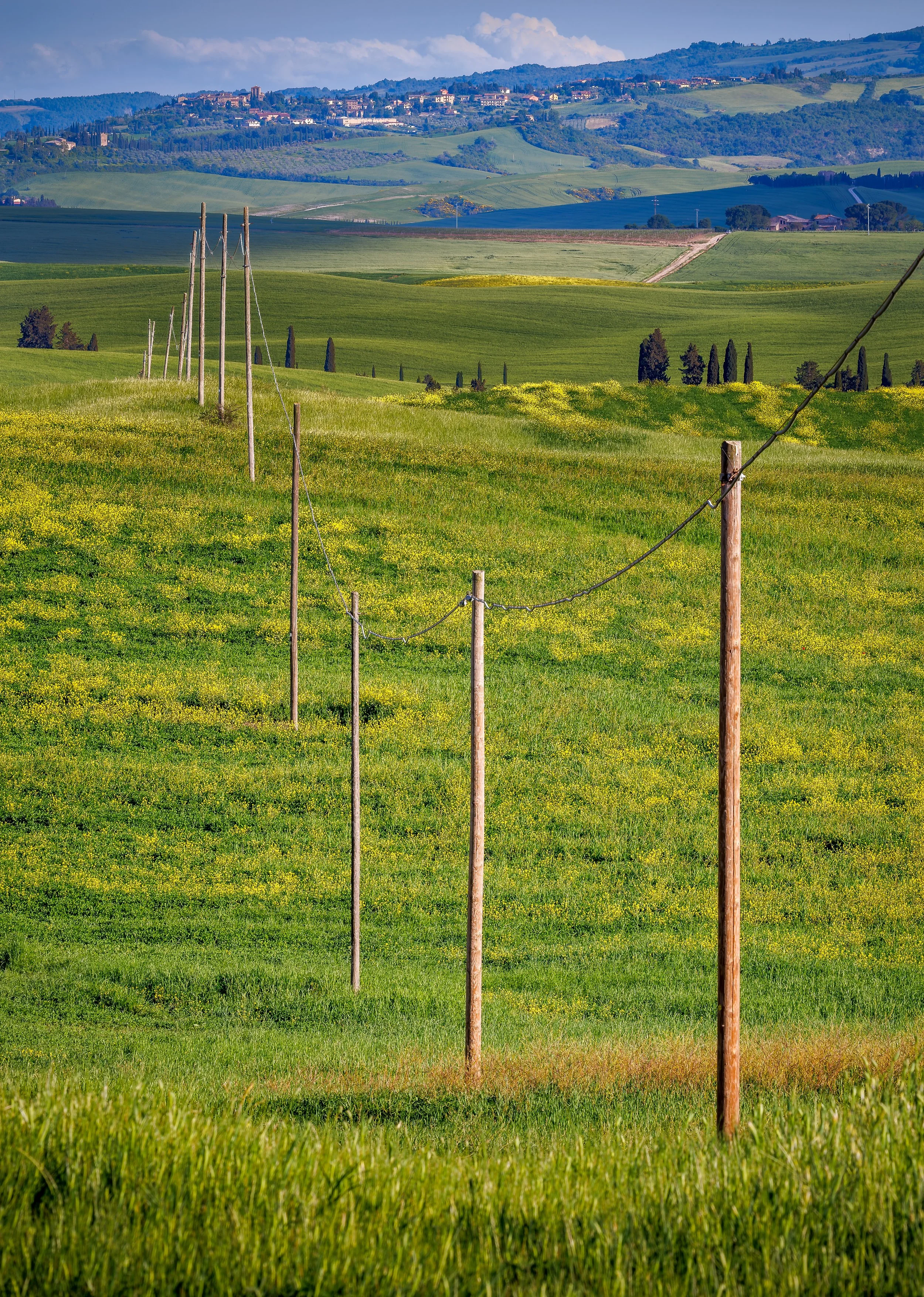 Rolling green hills with a wire fence and utility poles, with a distant village on a hillside in the background under a partly cloudy sky.