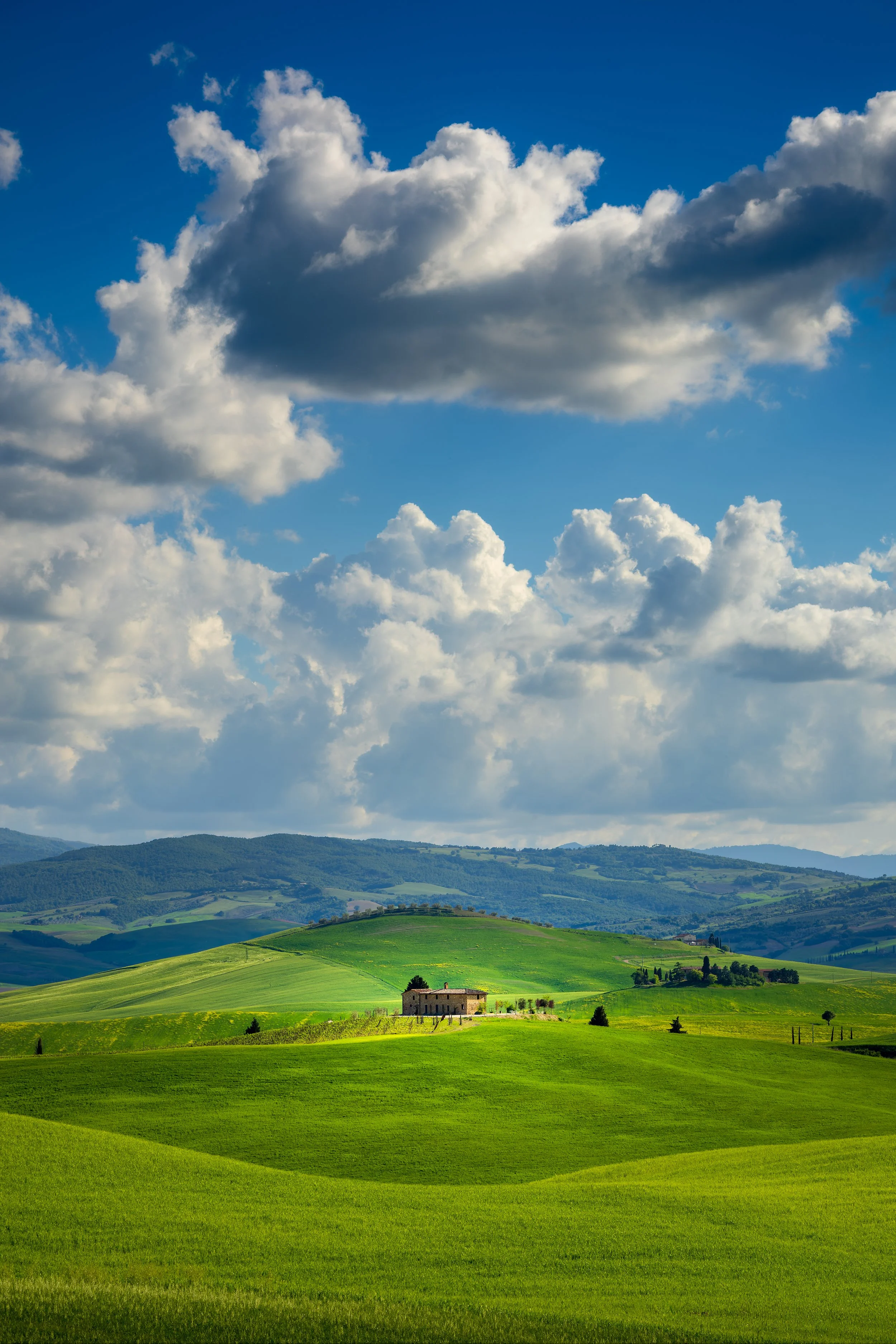 Green rolling hills with small stone houses, a distant mountain range, partly cloudy sky, and bright sunlight.
