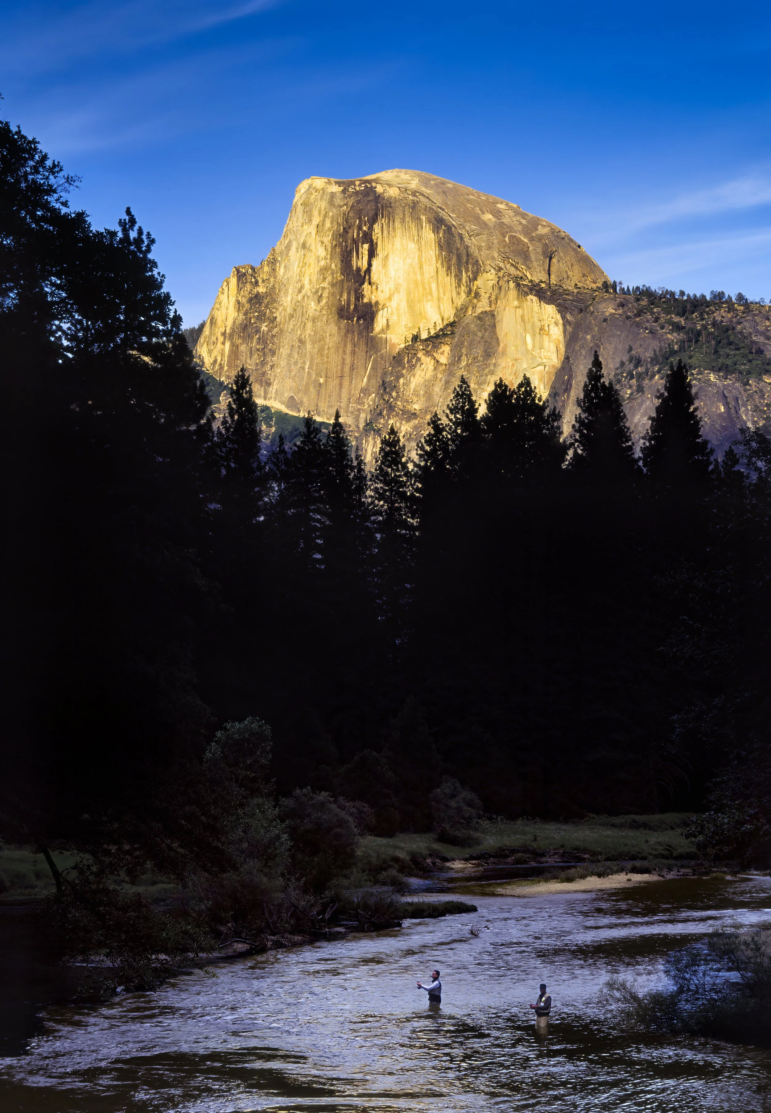 A large granite mountain, Half Dome, with a steep face, is illuminated by sunlight behind a dark forest. Two people are standing in a river, fishing, with trees and a mountain landscape in the background.