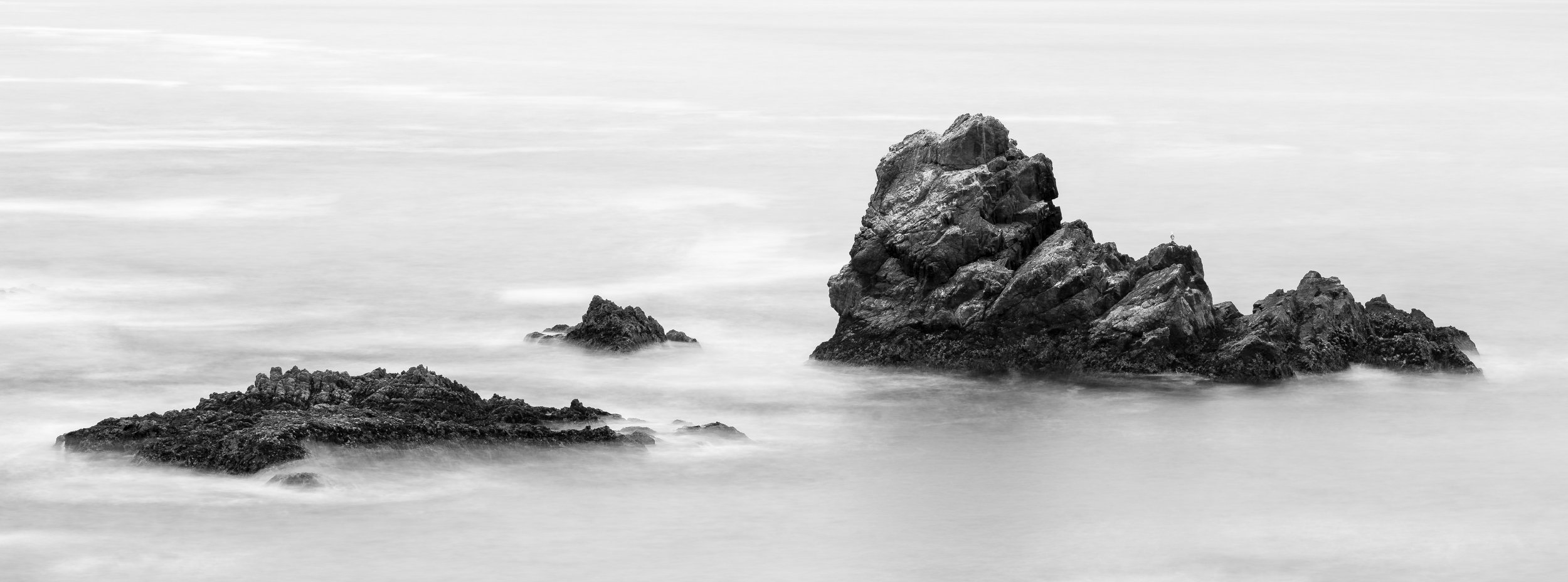 Black and white photo of rocks emerging from calm water, with a blurred effect created by long exposure.