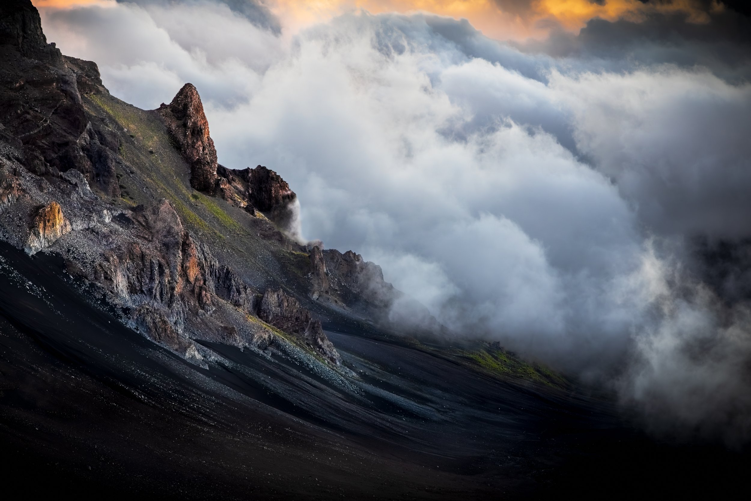A rugged mountain cliff with dark, rocky terrain and patches of green moss, partially obscured by a thick layer of fog or clouds, with a dramatic sky in the background.