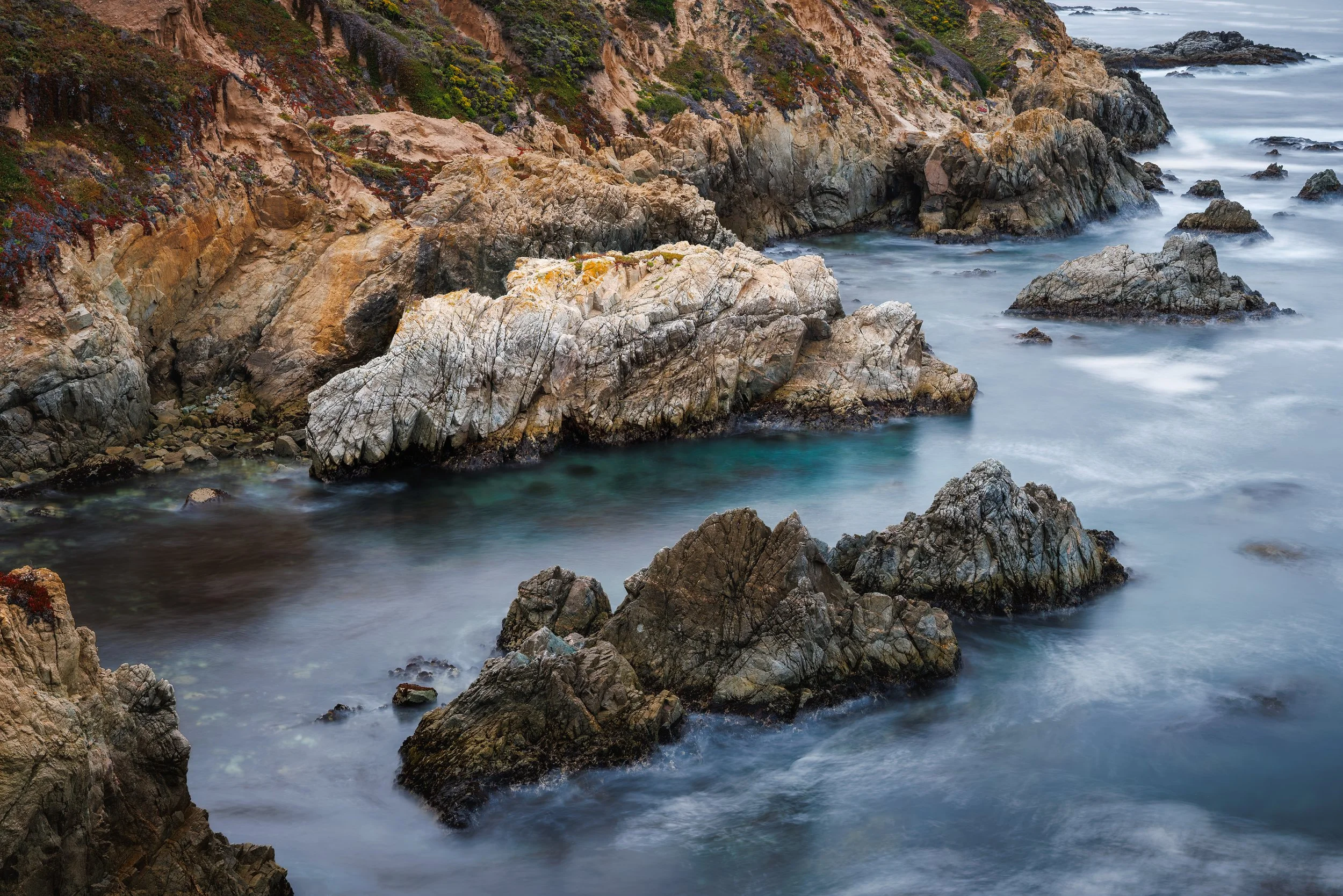 Rocky coastline with large rocks and cliffs, with waves crashing against them under a cloudy sky.