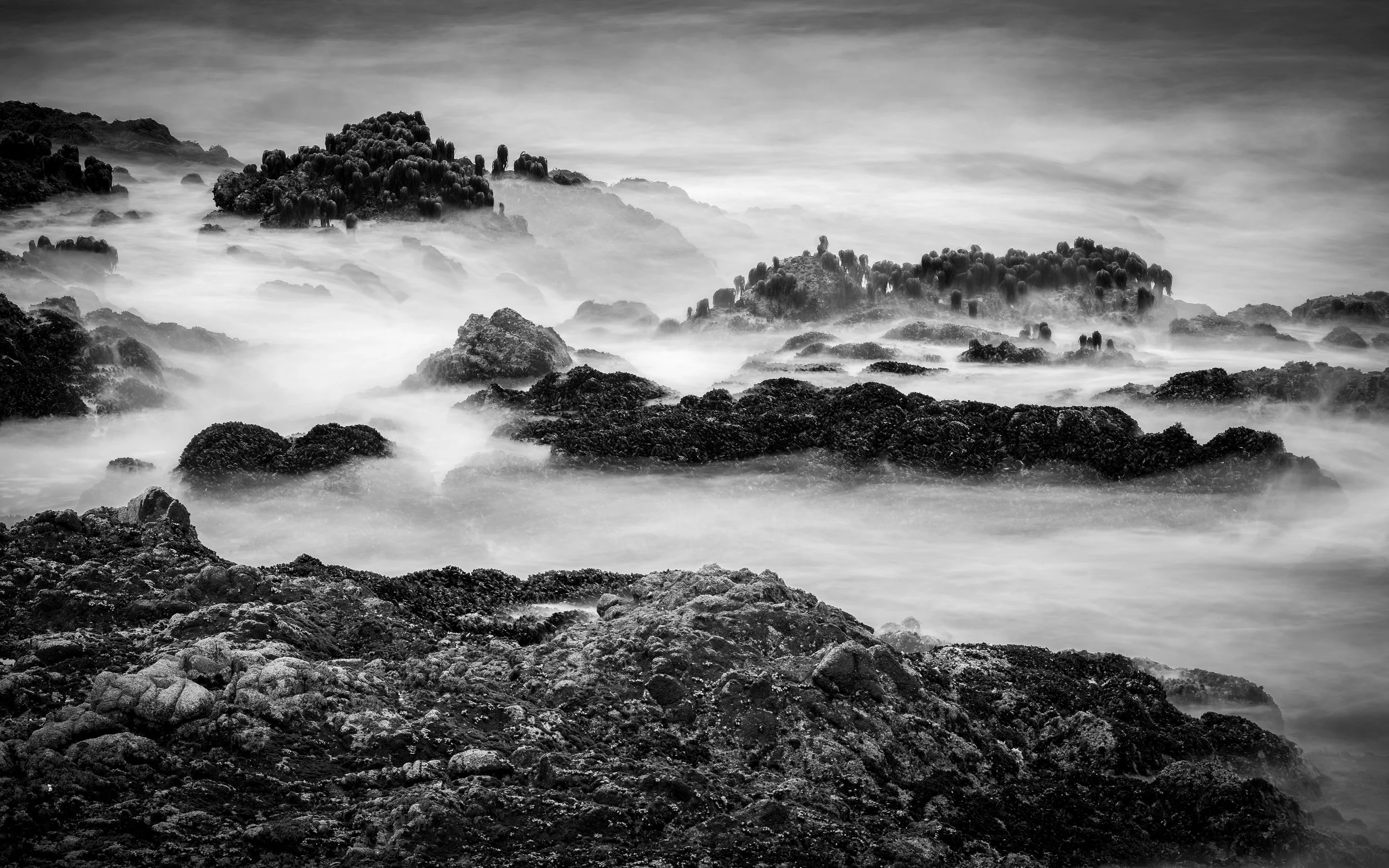 Black and white photograph of rocky coastal landscape with waves crashing against rocks and mist rising from the water.