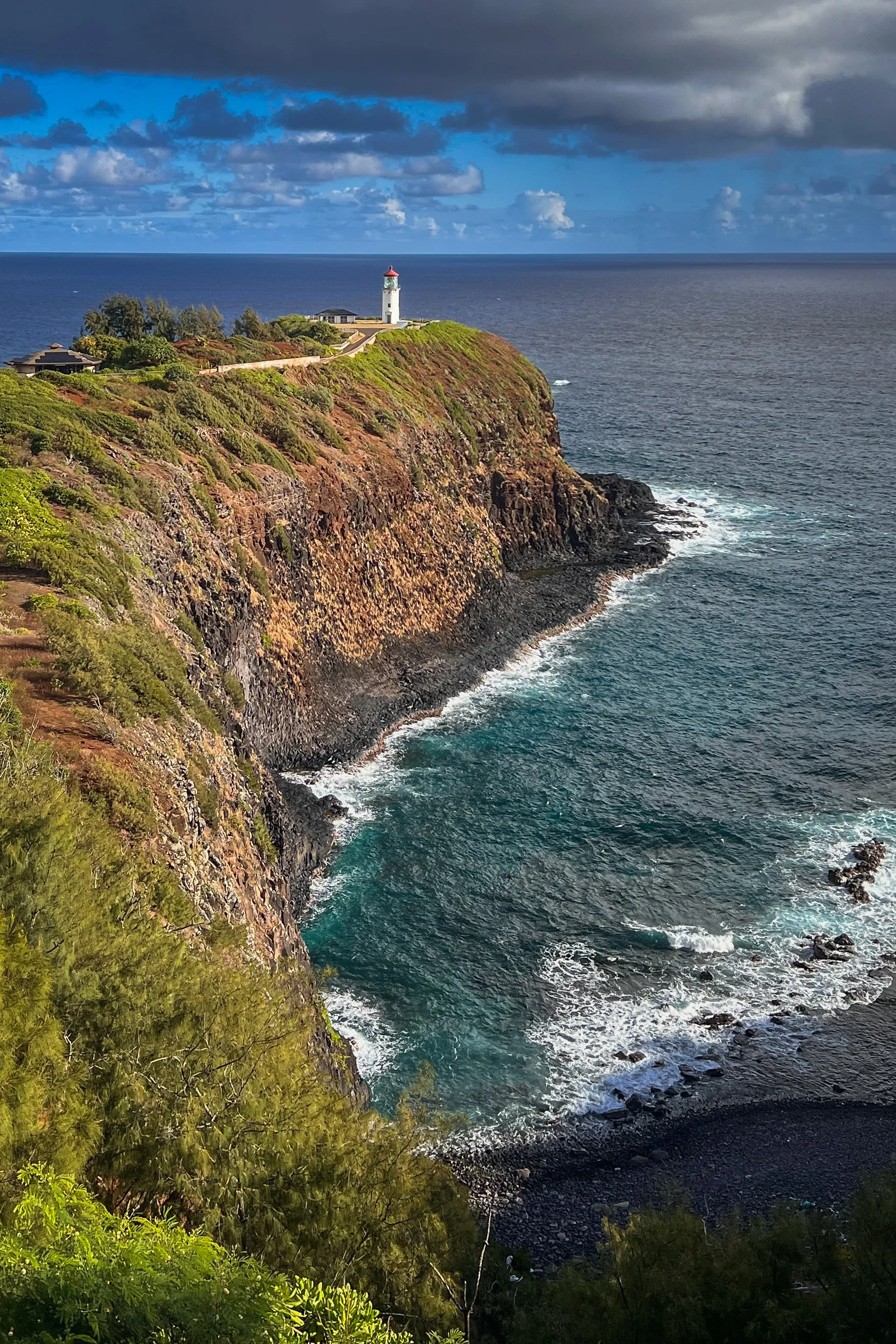 Kauai, Hawaii - A coastal scene with a lighthouse on a cliff, surrounded by some trees and houses. The ocean waves crash against the rocky shoreline, and dark clouds cover part of the sky.