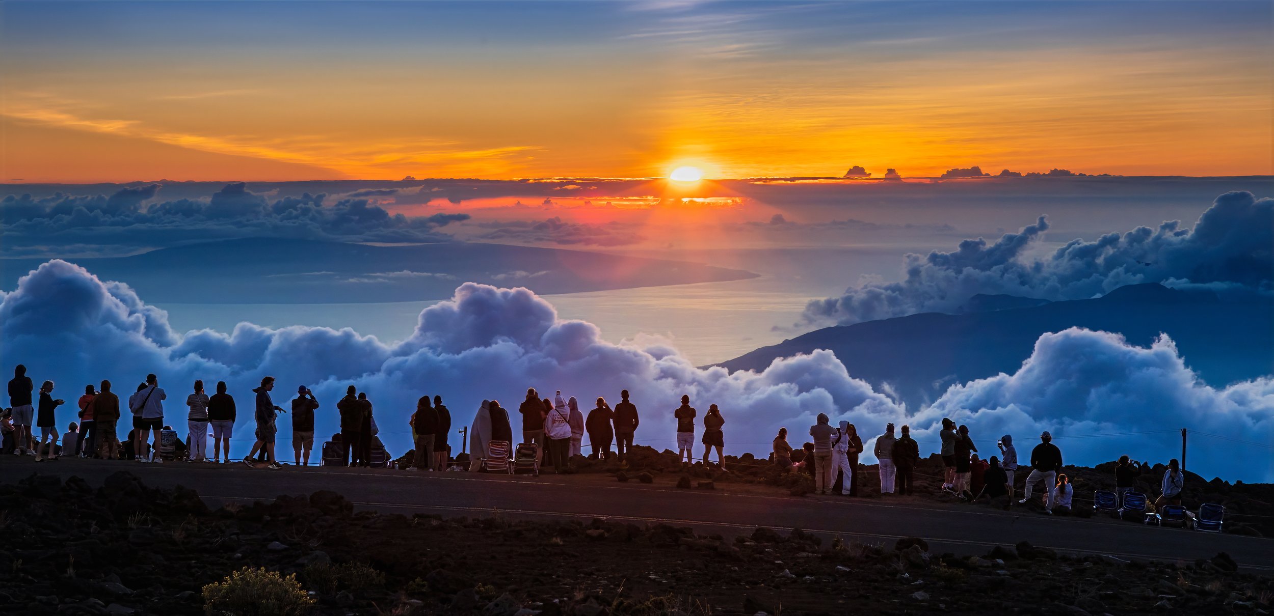 People standing on a mountain observing a sunset through clouds over mountains and water, with vibrant orange, yellow, and blue sky.