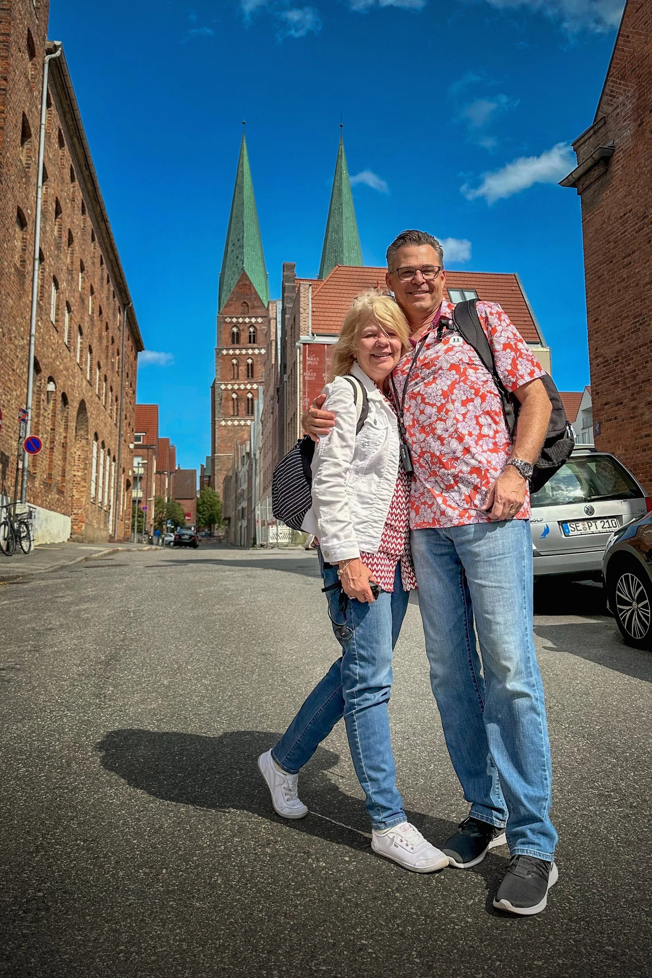 A man and a woman standing together on a city street, smiling. The man wears a red and white floral shirt, glasses, and has a backpack. The woman wears a white jacket, red and white patterned dress, and a backpack. Behind them are brick buildings and church steeples with a bright blue sky.