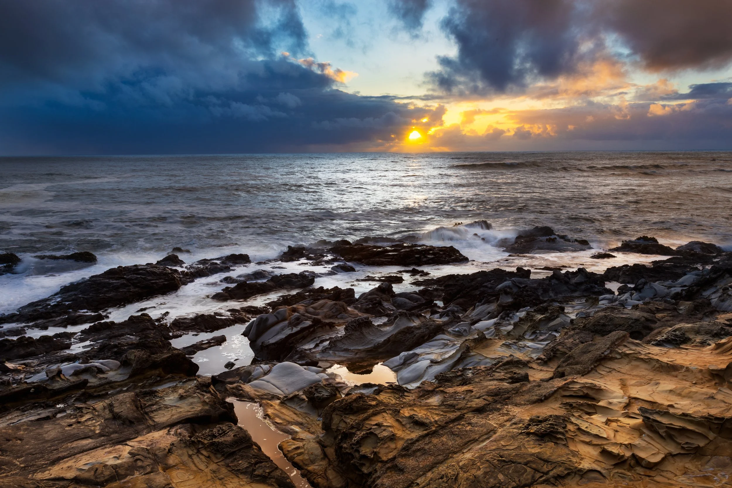 A sunset over the ocean with clouds and rocky shoreline.