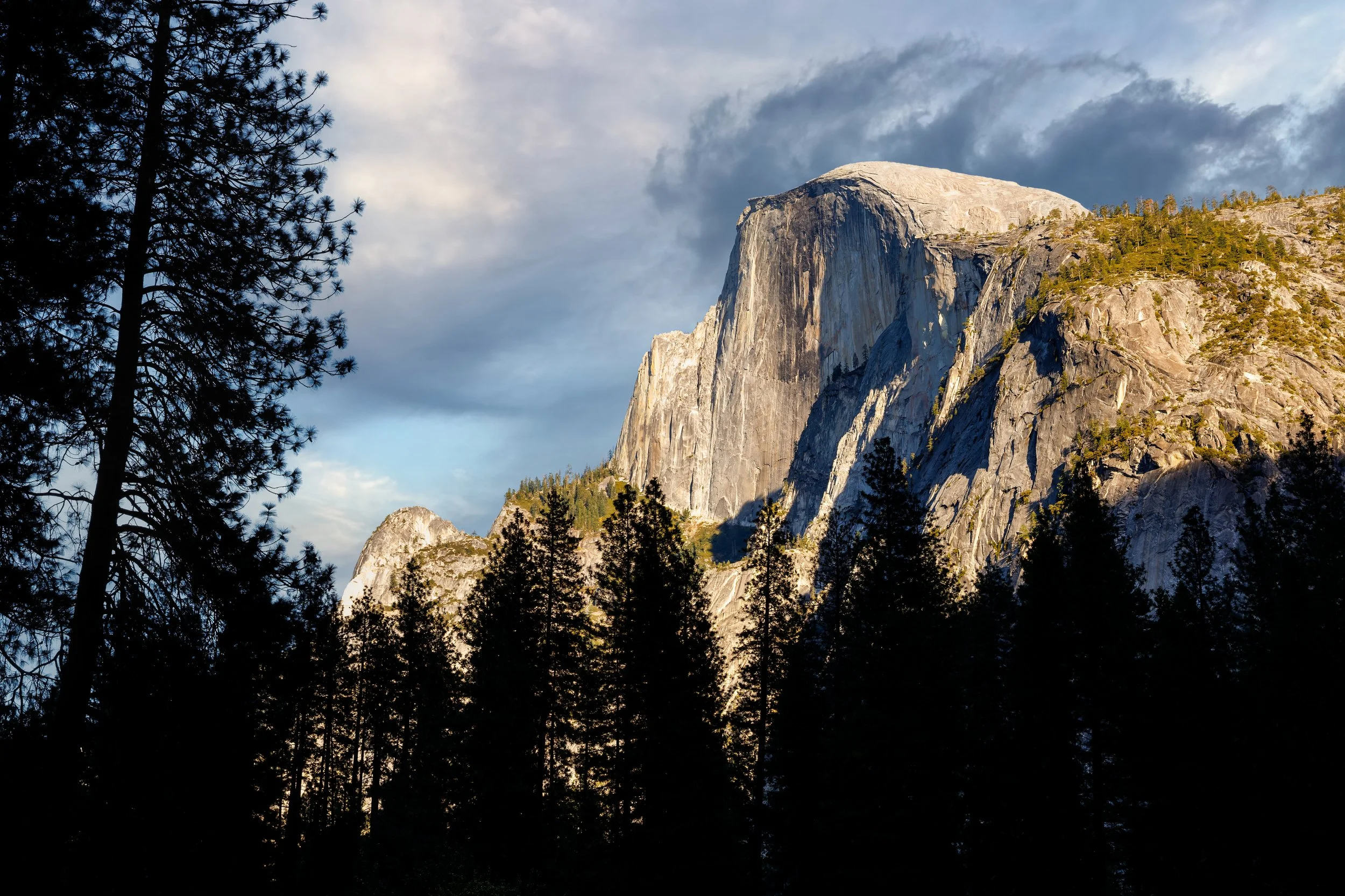 Mountain with a flat top, likely Half Dome in Yosemite National Park, surrounded by pine trees and a cloudy sky.