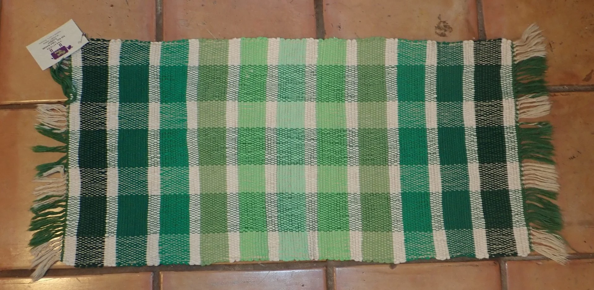 A rectangular woven table runner with a green, white, and black checkered pattern, fringed on both ends, lying on a tiled floor.
