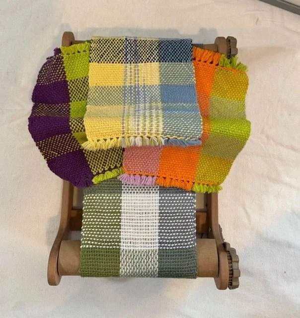 A miniature wooden loom with a colorful woven blanket draped over it. The blanket features various woven patterns and vibrant colors, including purple, green, yellow, pink, orange, and blue.