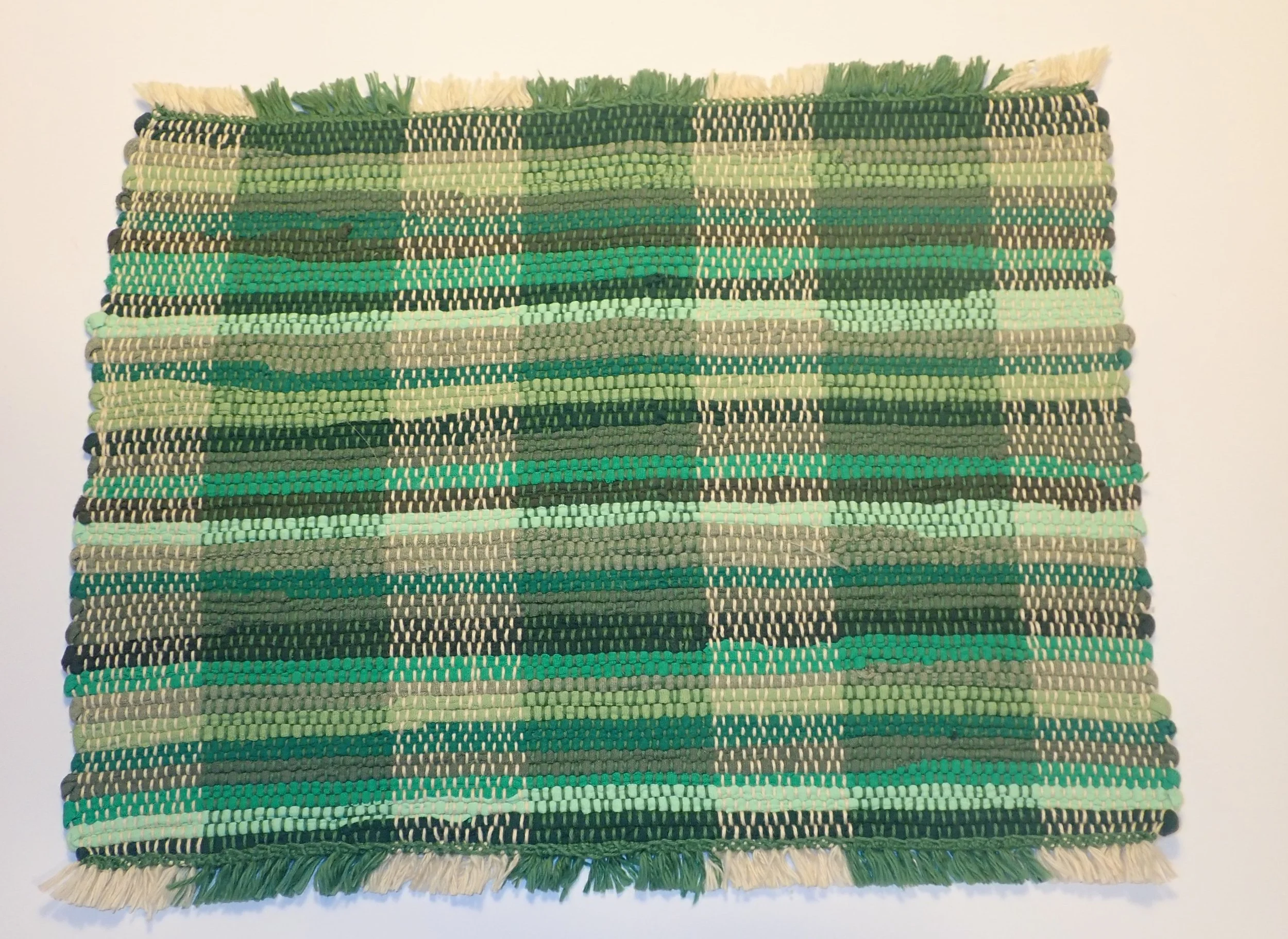 A rectangular woven rug with horizontal stripes in shades of green, black, and beige, featuring fringes on the shorter sides.