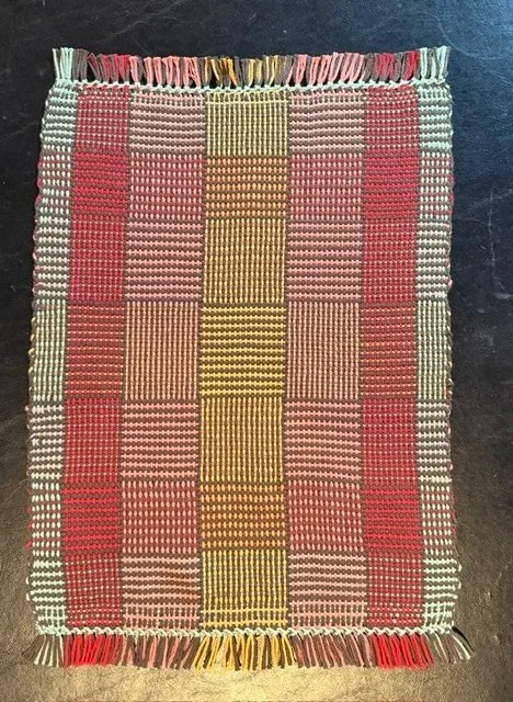 Colorful woven textile with a checkered pattern of red, pink, yellow, and white squares, fringed edges at the top and bottom.