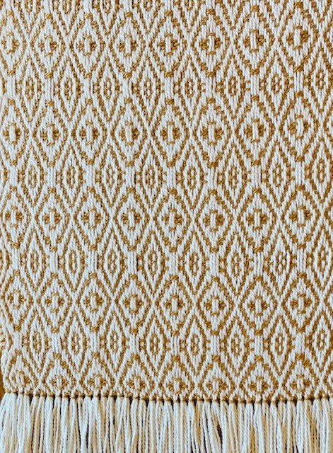 Close-up of a woven fabric with a repeating diamond and geometric pattern in beige and white, with a ruffled edge at the bottom.