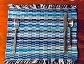 A woven fabric placemat with blue and white stripes, with silver utensils placed on top, on a wooden surface.