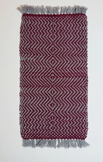 A small rectangular woven rug with red and white geometric patterns and fringes on both ends hanging against a plain wall.