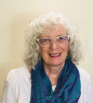An woman with white curly hair wearing glasses, a blue scarf, and a white top, smiling at the camera.