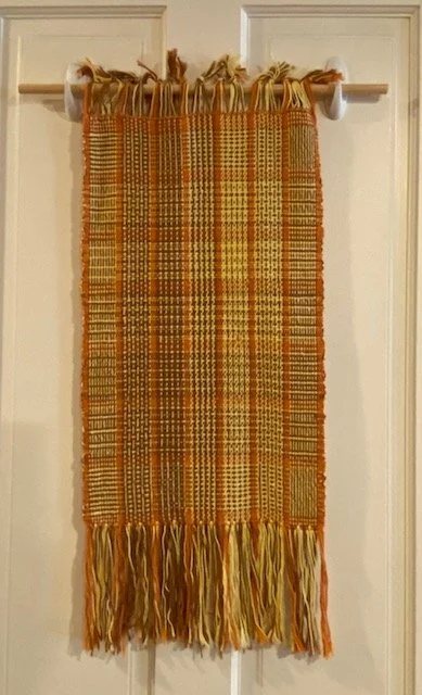 A decorative woven textile with a plaid pattern in orange, yellow, and green hanging on a curtain rod with white hooks in front of a white door.