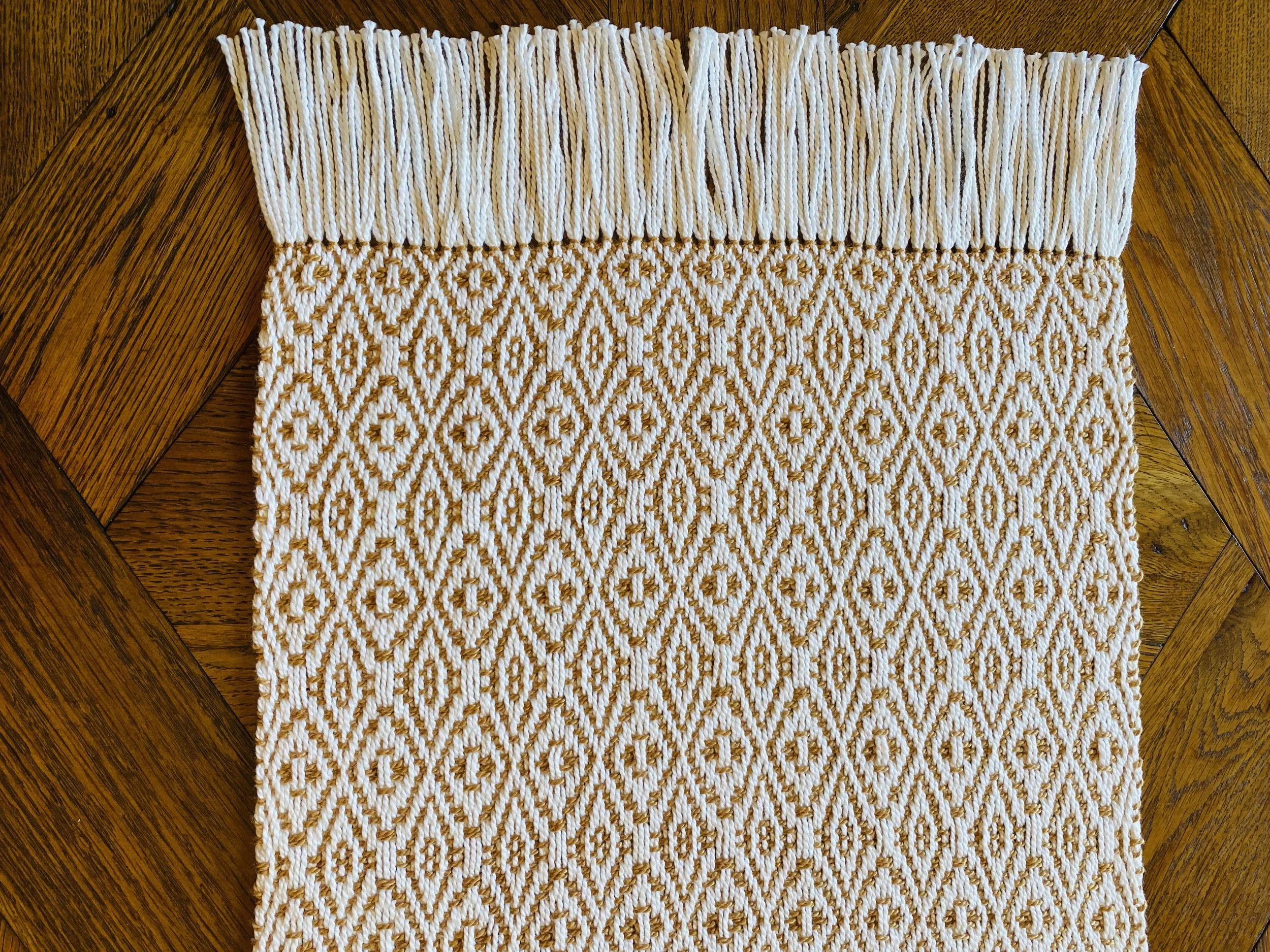 A woven textile with a white and gold patterned design, placed on a wooden surface.