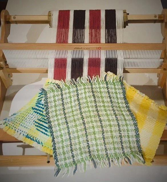 Colorful woven fabric or blanket draped over a wooden loom with partially woven fabric still on the loom. The fabric features various patterns and colors, including green, blue, yellow, and white.