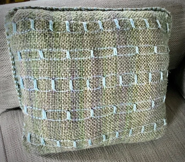 A decorative throw pillow with a textured, woven fabric featuring horizontal rows of looped embroidery stitches in light blue, placed on a beige fabric surface.