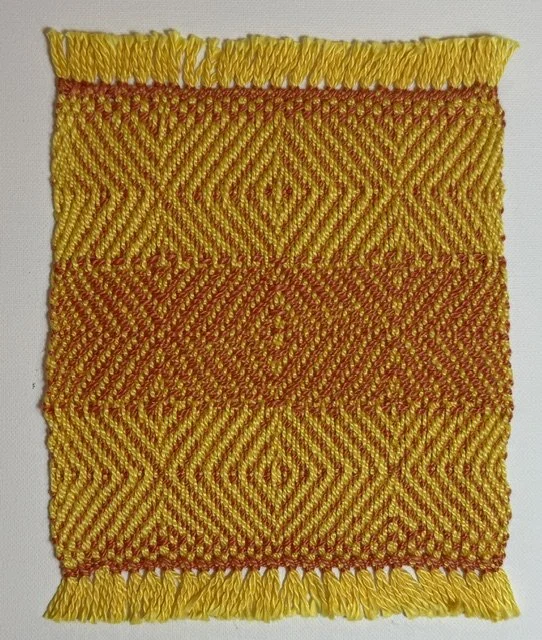 Close-up of a square woven textile with yellow and reddish-orange geometric patterns and fringed edges.