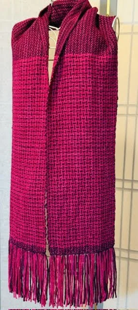 Pink and black woven scarf with fringe hanging on a wire grid