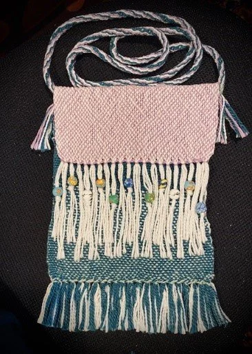 A woven shoulder bag with a pink upper section and a blue and white fringed bottom, decorated with small colored beads hanging from the fringes.