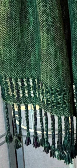 Close-up of a woven green textile with fringe at the bottom, displayed against a neutral background.