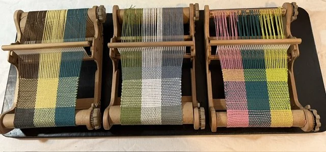 Three small wooden weaving looms with colorful woven fabric samples in yellow, green, blue, and pink shades.
