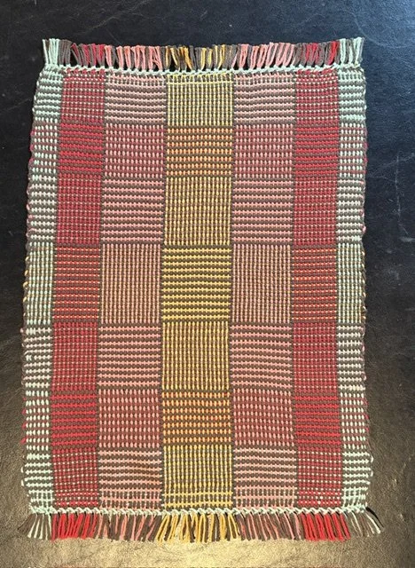 A woven textile rug with a checkered pattern of red, pink, yellow, and beige squares, fringed at the top and bottom edges.