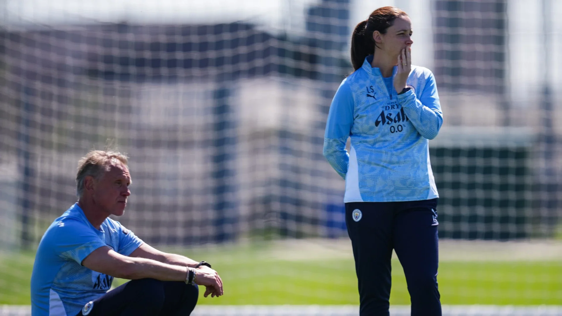 Two female soccer players in blue training jerseys on a field, one sitting and one standing near the goal net.