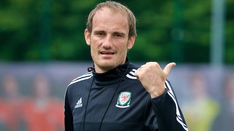 A man in a black sports jacket with the Welsh football badge, giving a thumbs-up gesture. Background is blurred greenery.